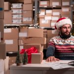 Weihnachtlich gekleideter Mitarbeiter sitzt im Lagerraum voller Kartons vor seinem Laptop