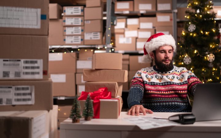 Weihnachtlich gekleideter Mitarbeiter sitzt im Lagerraum voller Kartons vor seinem Laptop